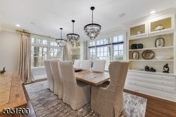 Dining Room featuring hardwood floors, baseboard molding, crown molding, built-in speakers, wall of triple windows with built-in window seat flanked by built-ins with open shelving and under storage, 3 chic chandeliers &recessed lights.