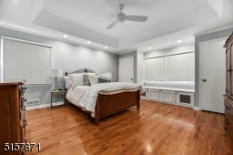Primary Bedroom Suite with hardwood floors, tray ceiling with recessed lights, ceiling fan, base and crown moldings, double window with a window seat and storage cabinets flanked by two fitted closets, an additional large fitted walk-in closet
