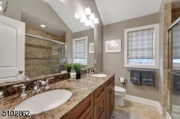 Primary Bathroom featuring stone floors, double vanity, two three light sconces and a glass enclosed stone tiled shower with built in bench